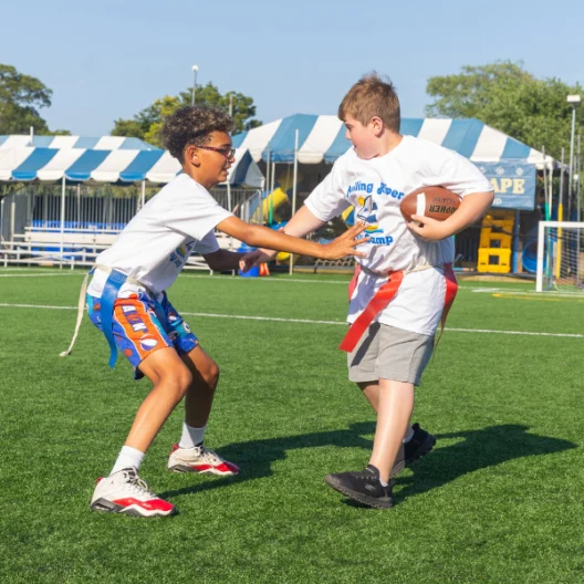 Two boys play flag football on a grass field; one boy reaches to grab a flag from the other, who is holding a football. Tents and equipment are visible in the background.