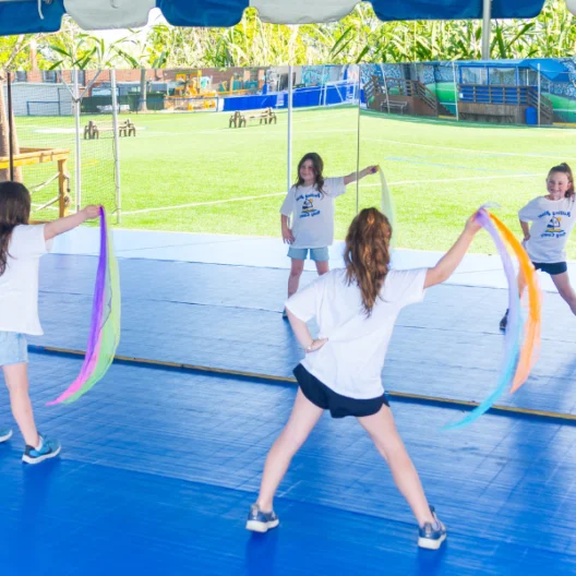 Four children in matching shirts practice a dance routine with colorful scarves inside a covered area next to an outdoor sports field.