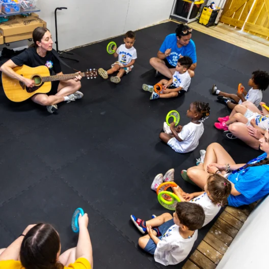 A group of children sit in a circle holding tambourines while an adult plays guitar; other adults supervise the activity in a classroom setting.