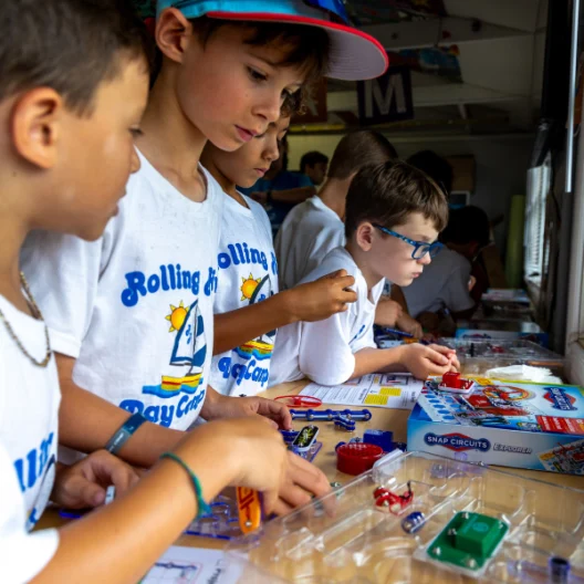 A group of children at a table assemble Snap Circuits kits while wearing matching Rolling River Day Camp t-shirts.