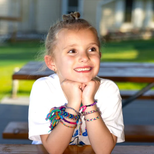 A young girl with braided bracelets on her wrists sits at a picnic table, resting her chin on her hands and smiling, with a blurred lawn and building in the background.