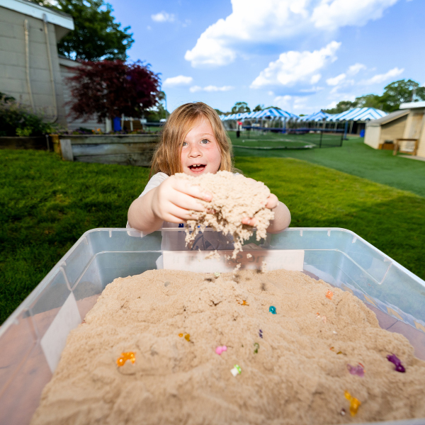 A young child plays with sand in a clear plastic bin outdoors, holding up a handful of sand with colorful objects mixed in. Tents and grass are visible in the background.