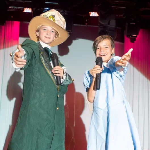Two children stand on stage under bright lights, holding microphones and reaching out with their free hands; one wears a green coat and hat, the other a light blue dress.