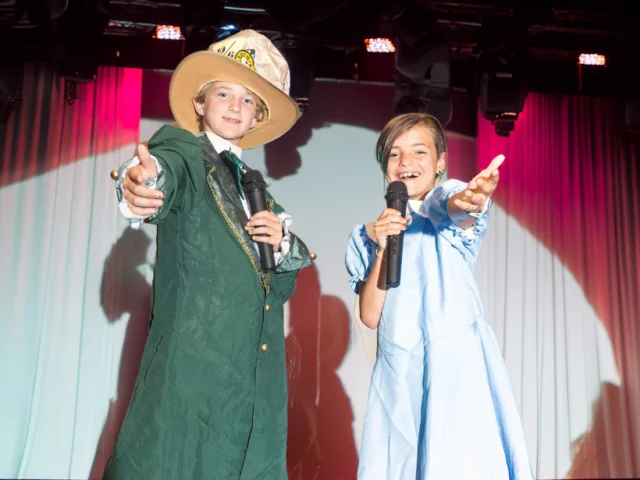 Two children stand on stage under bright lights, holding microphones and reaching out with their free hands; one wears a green coat and hat, the other a light blue dress.