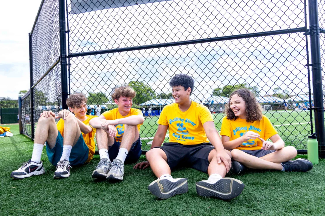 Four teenagers in matching yellow shirts sit on grass and smile together by a chain-link fence at an outdoor sports field.