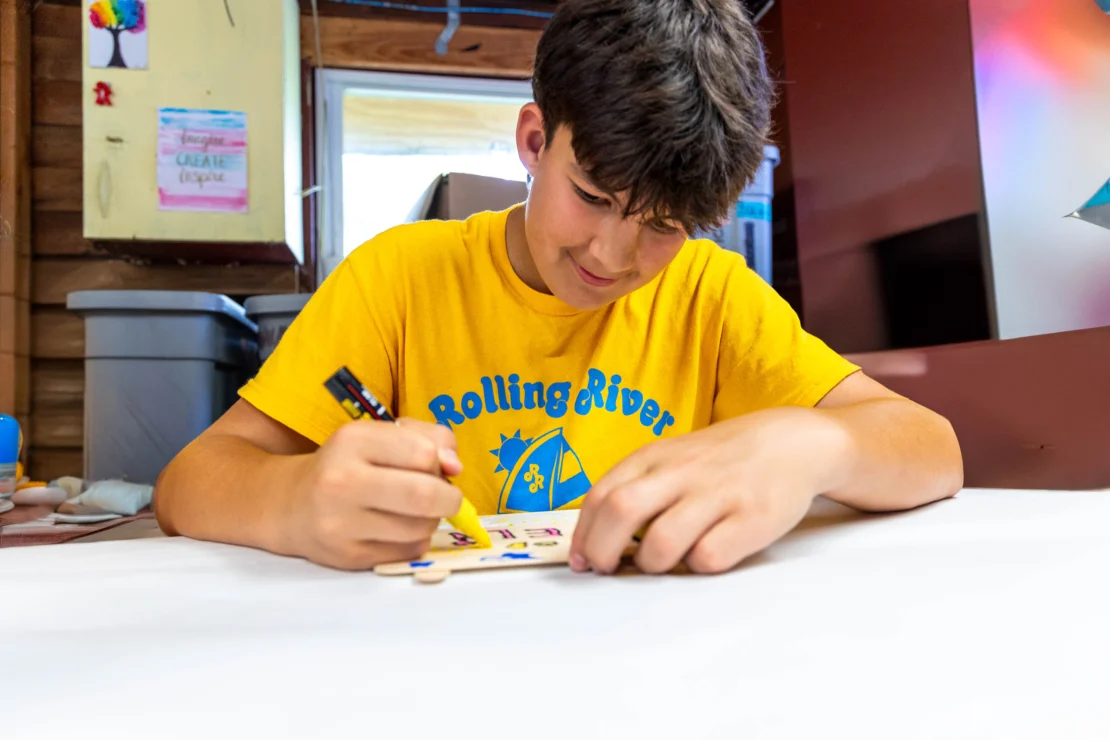A boy in a yellow "Rolling River" camp T-shirt colors a wooden craft project at a table indoors.