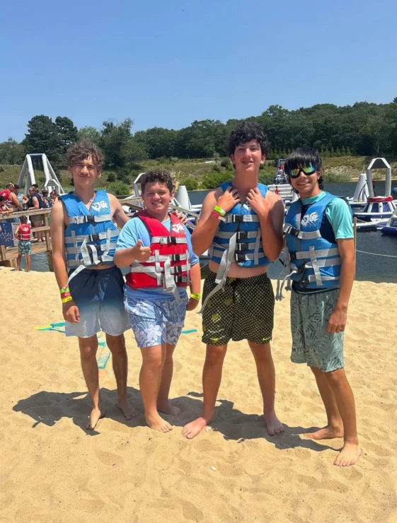 Four young people wearing life jackets and swimwear stand on a sandy beach, posing for a photo with boats and water in the background on a sunny day.