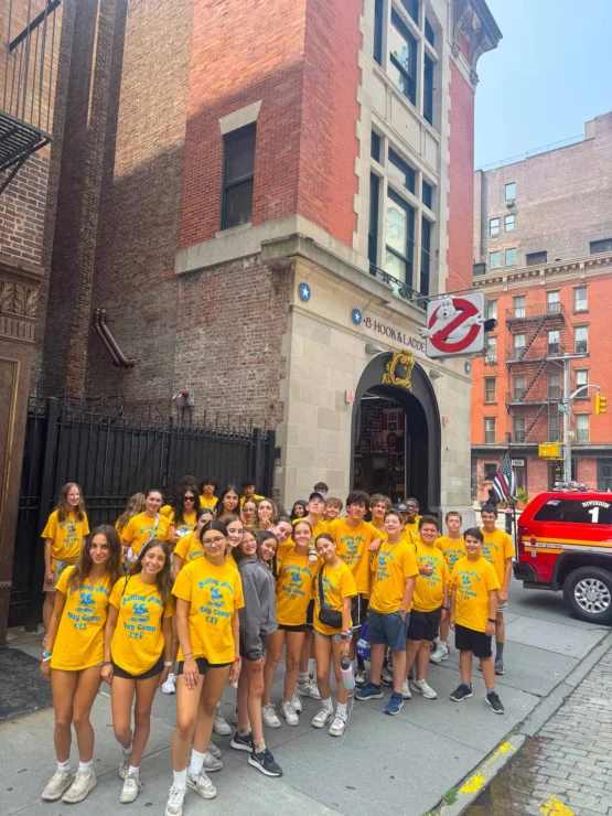 A group of teenagers in matching yellow shirts pose in front of the Hook & Ladder 8 firehouse, known for its Ghostbusters sign, in an urban setting.