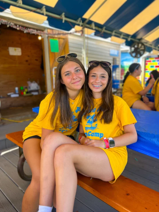 Two young women wearing matching yellow camp T-shirts sit closely together on a bench under a striped canopy, smiling at the camera.