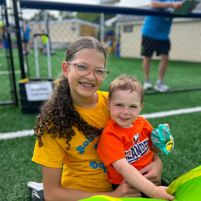 A girl with glasses and braces sits on artificial turf, smiling and holding a young boy in an orange shirt who is holding a snack. They are outdoors near a fence.