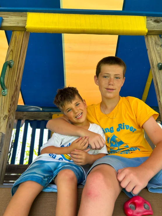 Two boys sit side by side on a wooden play structure, smiling, with one boy’s arm around the other. Both wear blue shorts and yellow or white T-shirts with matching graphics.