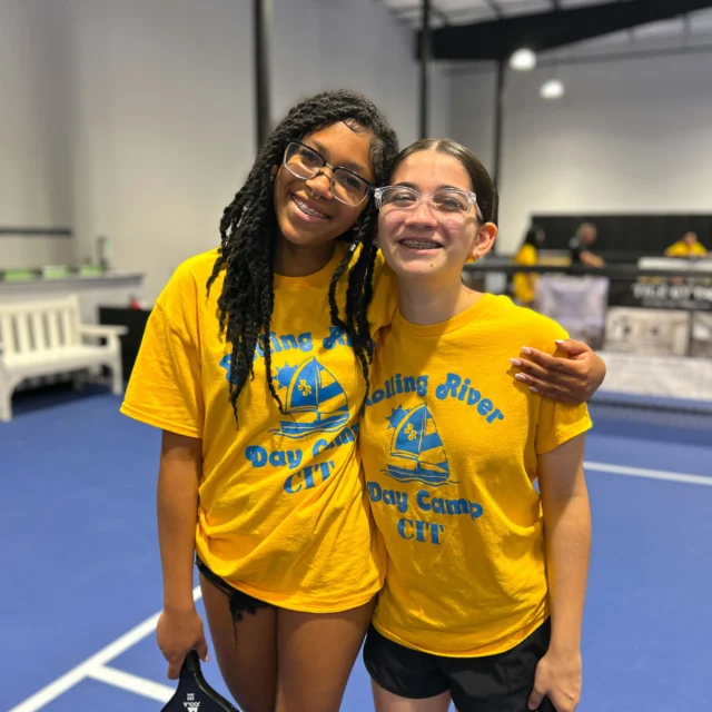 Two girls in yellow "Rolling River Day Camp CIT" shirts stand and smile on an indoor court, holding pickleball paddles.
