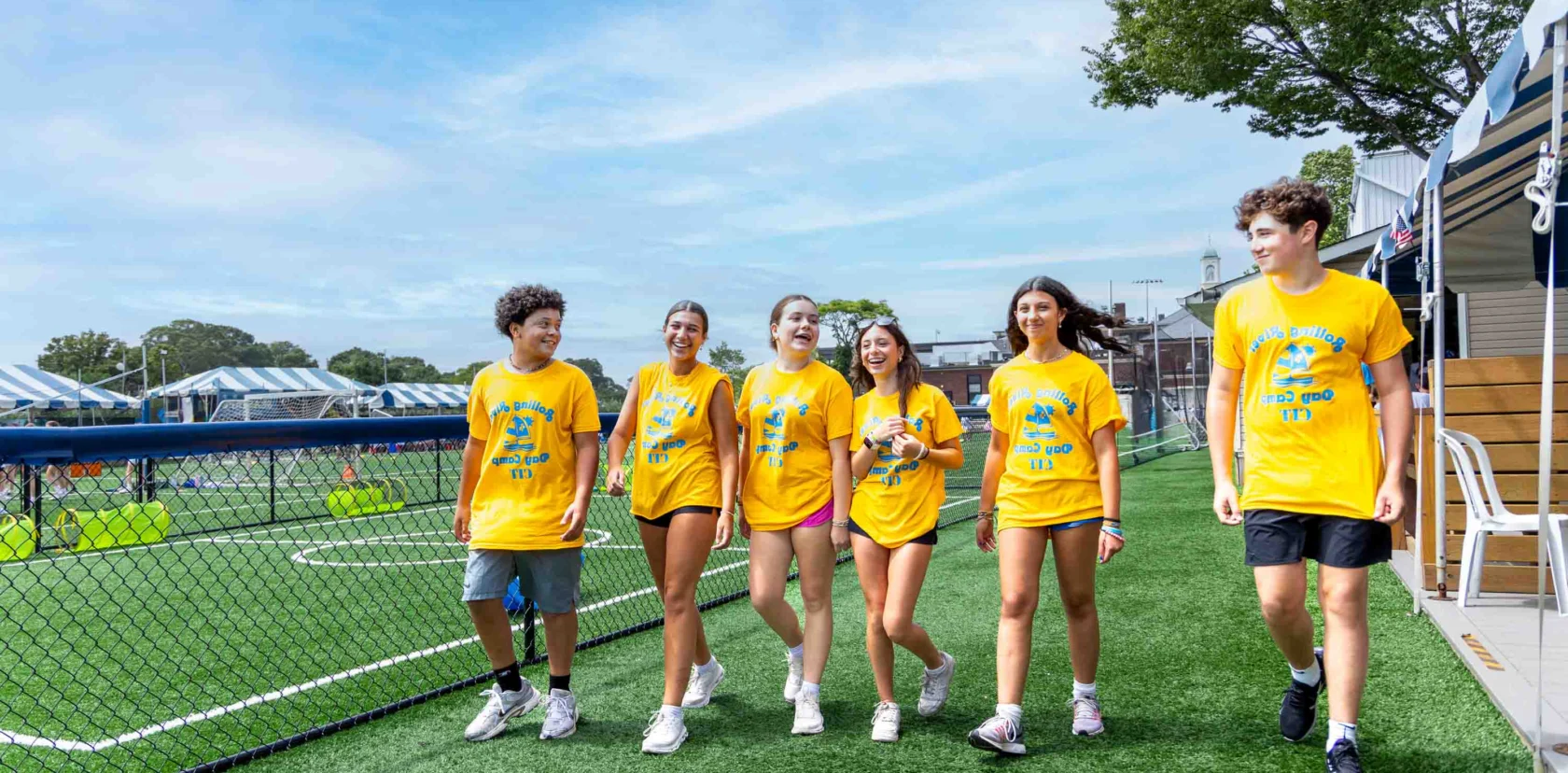 Six teenagers wearing matching yellow shirts walk together on artificial turf near a chain-link fence, with trees and white tents visible in the background.