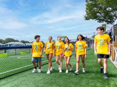 Six teenagers wearing matching yellow shirts walk together on artificial turf near a chain-link fence, with trees and white tents visible in the background.