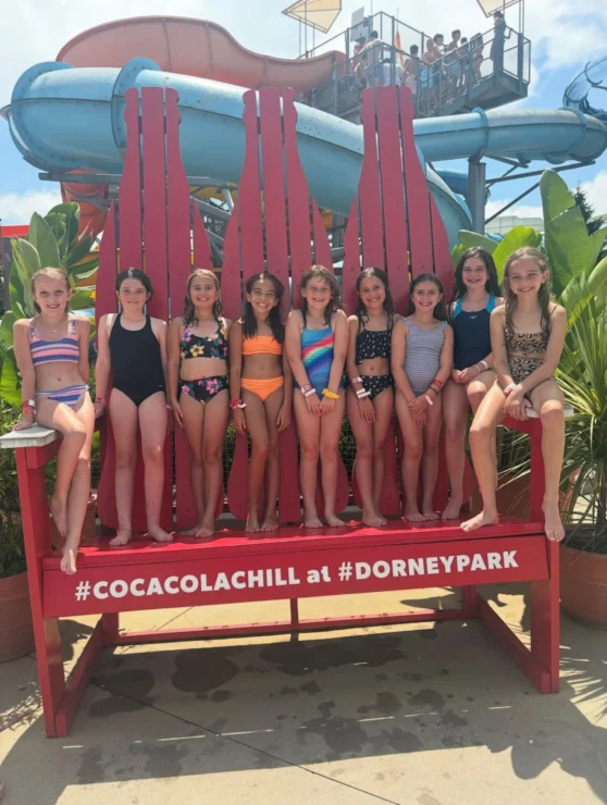 Nine girls in swimsuits sit and stand on a large red bench shaped like Coca-Cola bottles at Dorney Park, with a water slide and plants in the background.