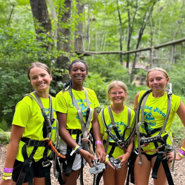 Four girls wearing harnesses and matching yellow T-shirts stand outdoors in a wooded area, smiling at the camera, likely preparing for a zip-lining or ropes course activity.