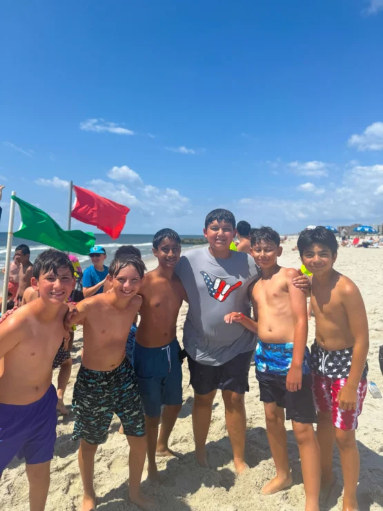 A group of six boys in swimsuits pose together on a sandy beach with red and green flags, under a sunny blue sky.