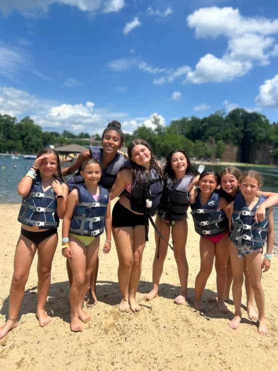 Seven girls wearing life jackets stand together and smile on a sandy beach by the water, with trees and a blue sky in the background.