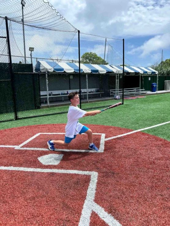 A boy in athletic clothes swings a baseball bat while kneeling at home plate on an outdoor baseball field.