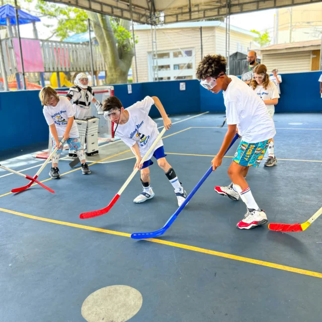 Four children play floor hockey indoors wearing white shirts and safety goggles. One child is goalie, while three compete for the ball with blue and red sticks. An adult supervises in the background.