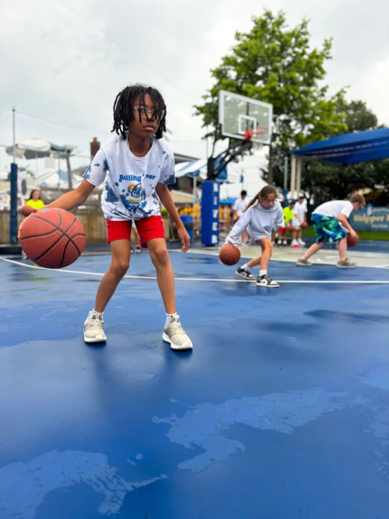 Children practice dribbling basketballs on an outdoor blue court, with one child in the foreground and others in the background.