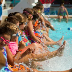 A group of children wearing swimsuits and goggles sit at the edge of a pool, splashing water with their feet.
