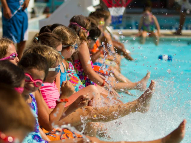 A group of children wearing swimsuits and goggles sit at the edge of a pool, splashing water with their feet.