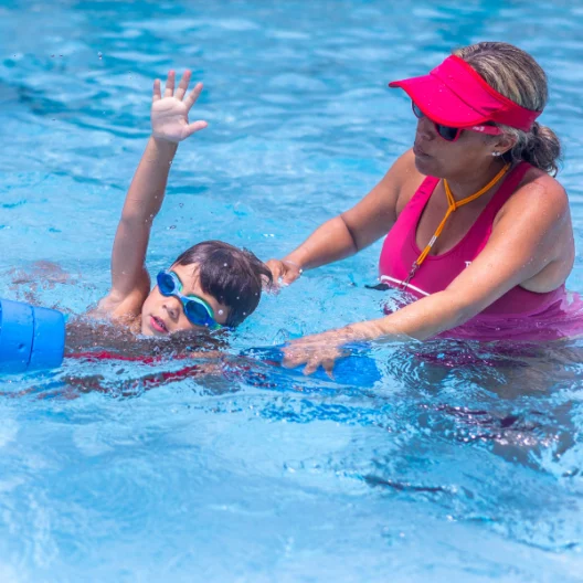A swim instructor in a red visor assists a young boy wearing blue goggles as he practices swimming in a pool with a blue kickboard.