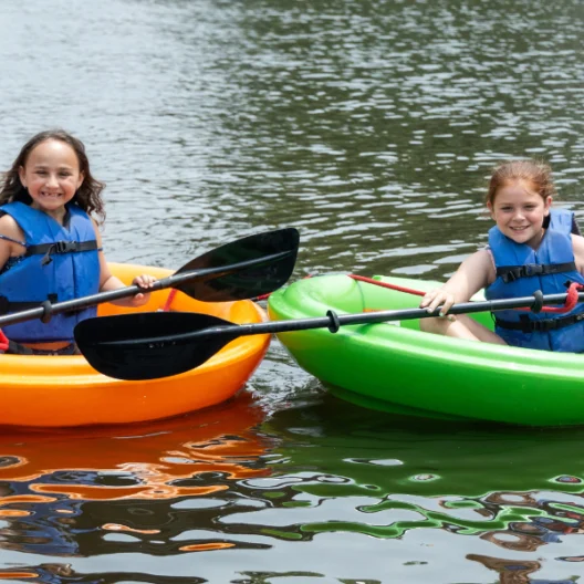 Two young girls in life jackets sit in orange and green kayaks on a calm body of water, holding paddles and smiling at the camera.