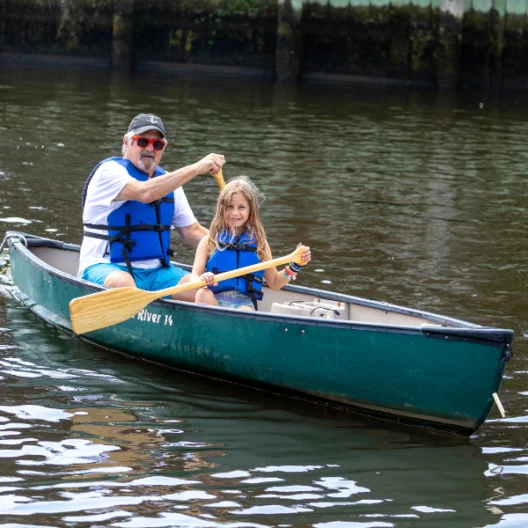 An older man and a young girl wearing life jackets paddle a green canoe together on a calm river.