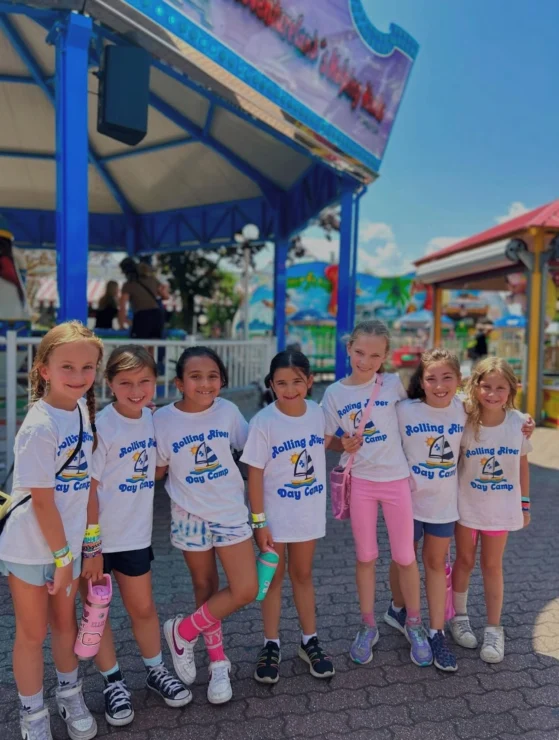 Seven young girls wearing matching "Rolling River Day Camp" shirts stand together and smile in front of an amusement park ride on a sunny day.