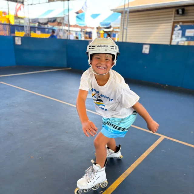 Smiling child wearing a helmet rollerblades indoors on a blue court, wearing a white t-shirt and blue shorts.