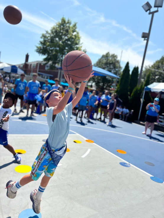 A young boy in colorful shorts jumps to shoot a basketball on an outdoor court, with spectators and other children in the background.