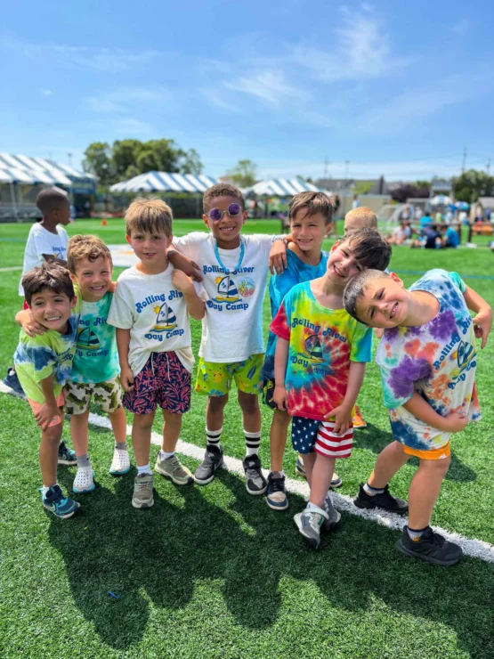 A group of young boys at a summer day camp stand together on a grassy field, smiling and wearing colorful tie-dye shirts under a sunny sky.