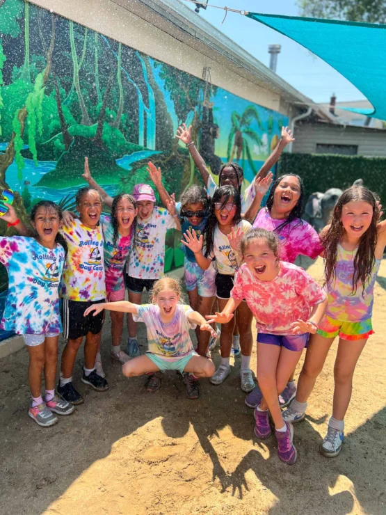 A group of children in colorful tie-dye shirts smile and pose outside, appearing wet and joyful, in front of a painted mural wall on a sunny day.