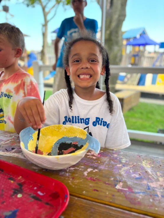 A smiling child sits at a painted wooden table holding a paintbrush over a colorful ceramic bowl, wearing a “Rolling River” t-shirt at an outdoor event.