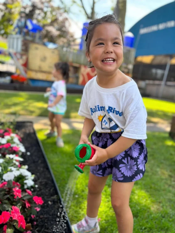 A young girl waters flowers with a small green watering can, smiling, while another child stands in the background on a sunny day.