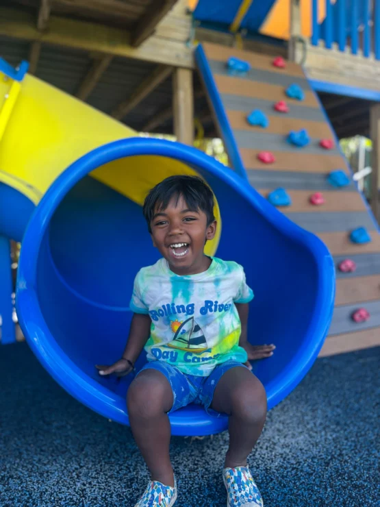 A young child sits smiling at the end of a blue slide on a playground, wearing a tie-dye "Rolling River Day Camp" T-shirt and shorts.