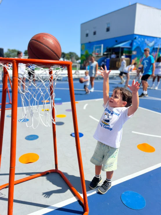 A young child attempts to shoot a basketball into a low hoop on an outdoor court, surrounded by other children and adults.