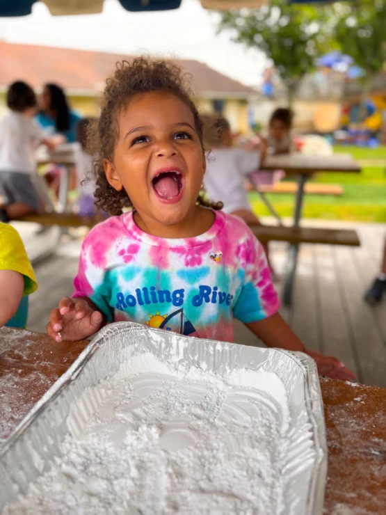 A young child wearing a tie-dye "Rolling River" shirt smiles and laughs while sitting at a table with a tray of white powder, outdoors with other children in the background.