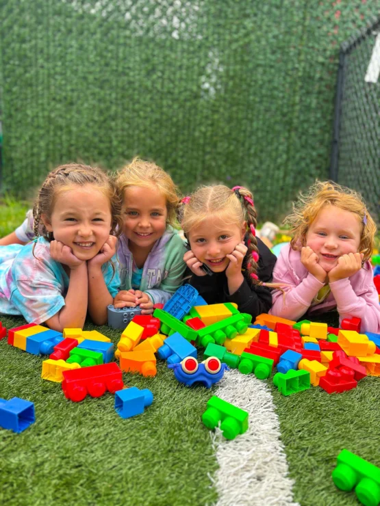 Four young children lie on artificial grass, smiling at the camera, with colorful plastic building blocks scattered in front of them.