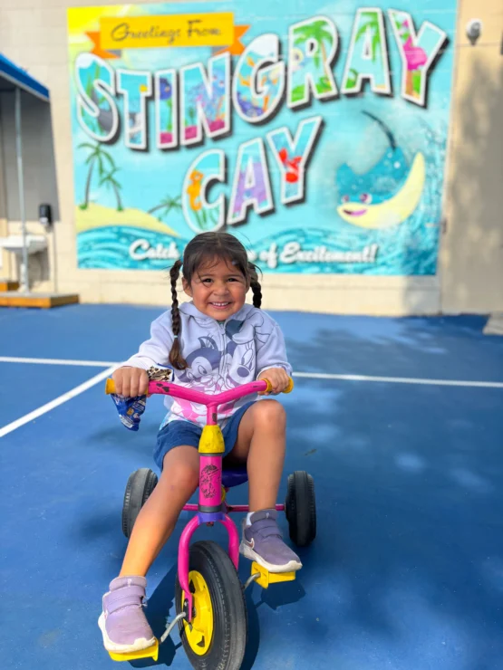 Young girl with pigtails smiles while riding a tricycle on a blue court in front of a colorful "Stingray Cay" mural.
