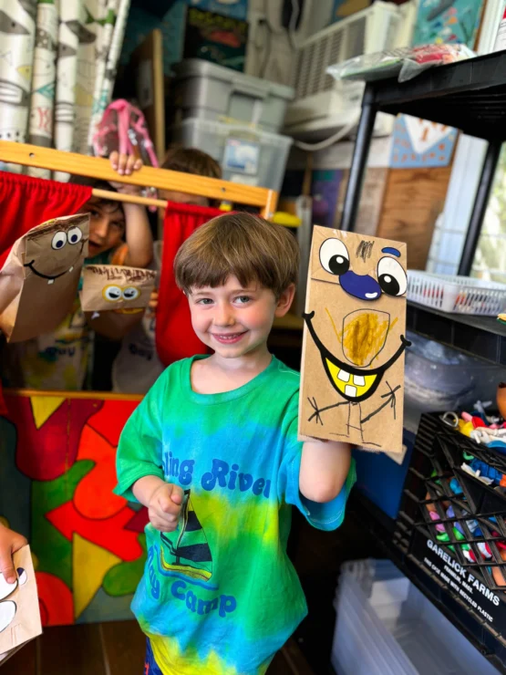 A young boy at an art camp holds a decorated paper bag puppet and smiles, with other children and colorful art supplies visible in the background.