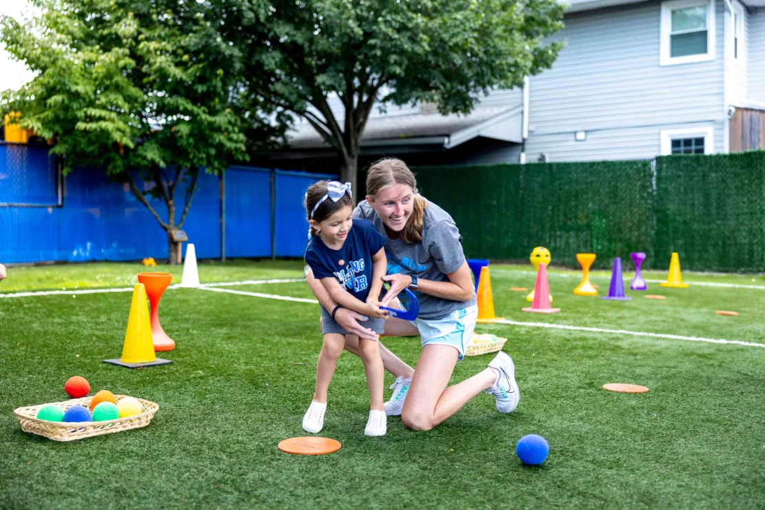 An adult and a child play together on a grassy field with colorful cones, balls, and baskets during an outdoor activity.