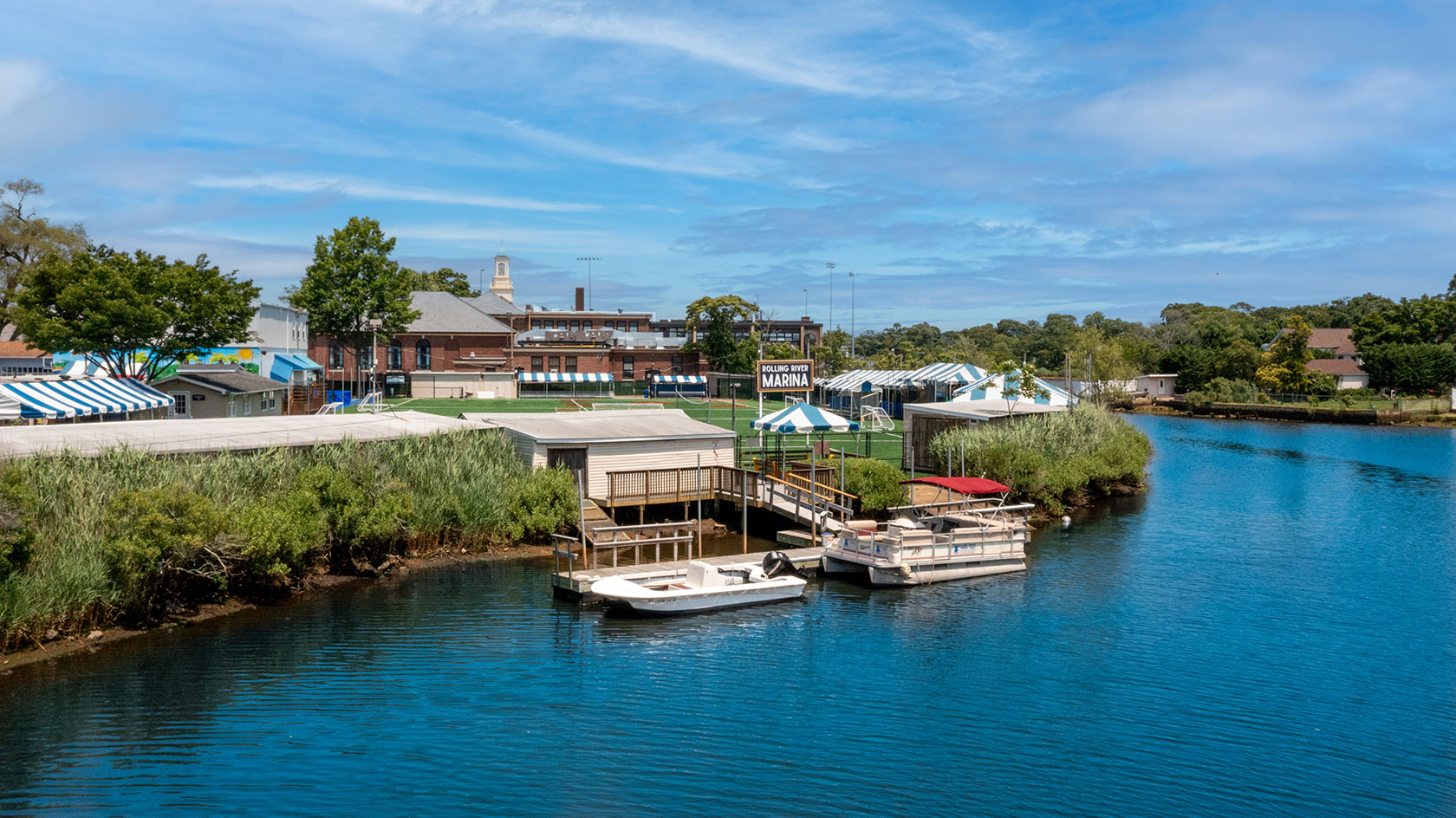 A small marina with boats docked along a calm river, surrounded by greenery and buildings under a blue sky.