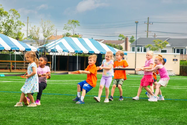 A group of young children play tug-of-war on a grassy field, with some wearing orange shirts and others in pastel colors, under a cloudy sky.