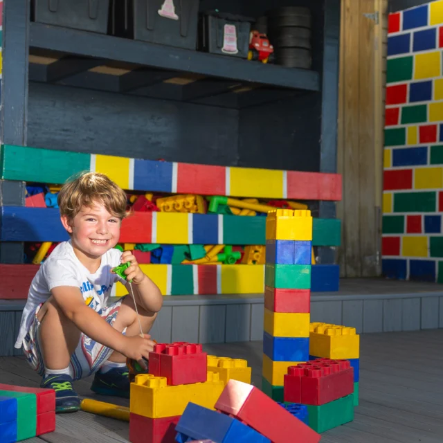 Young boy sitting on the floor inside a play area, smiling while building a tower with large colorful toy blocks.