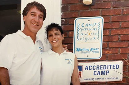 Two adults in matching white shirts stand smiling in front of a brick wall with camp signage, including “Accredited Camp” and “Camp gives kids a world of good.”.