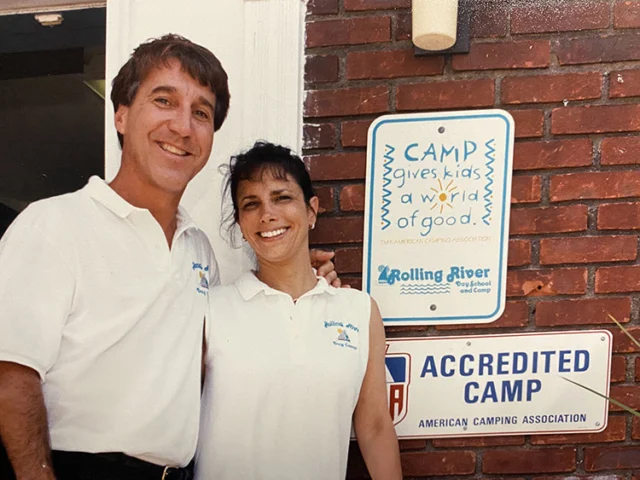 Two adults in matching white shirts stand smiling in front of a brick wall with camp signage, including “Accredited Camp” and “Camp gives kids a world of good.”.