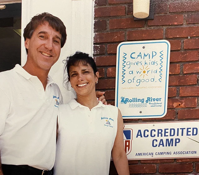 Two adults in matching white shirts stand smiling in front of a brick wall with camp signage, including “Accredited Camp” and “Camp gives kids a world of good.”.